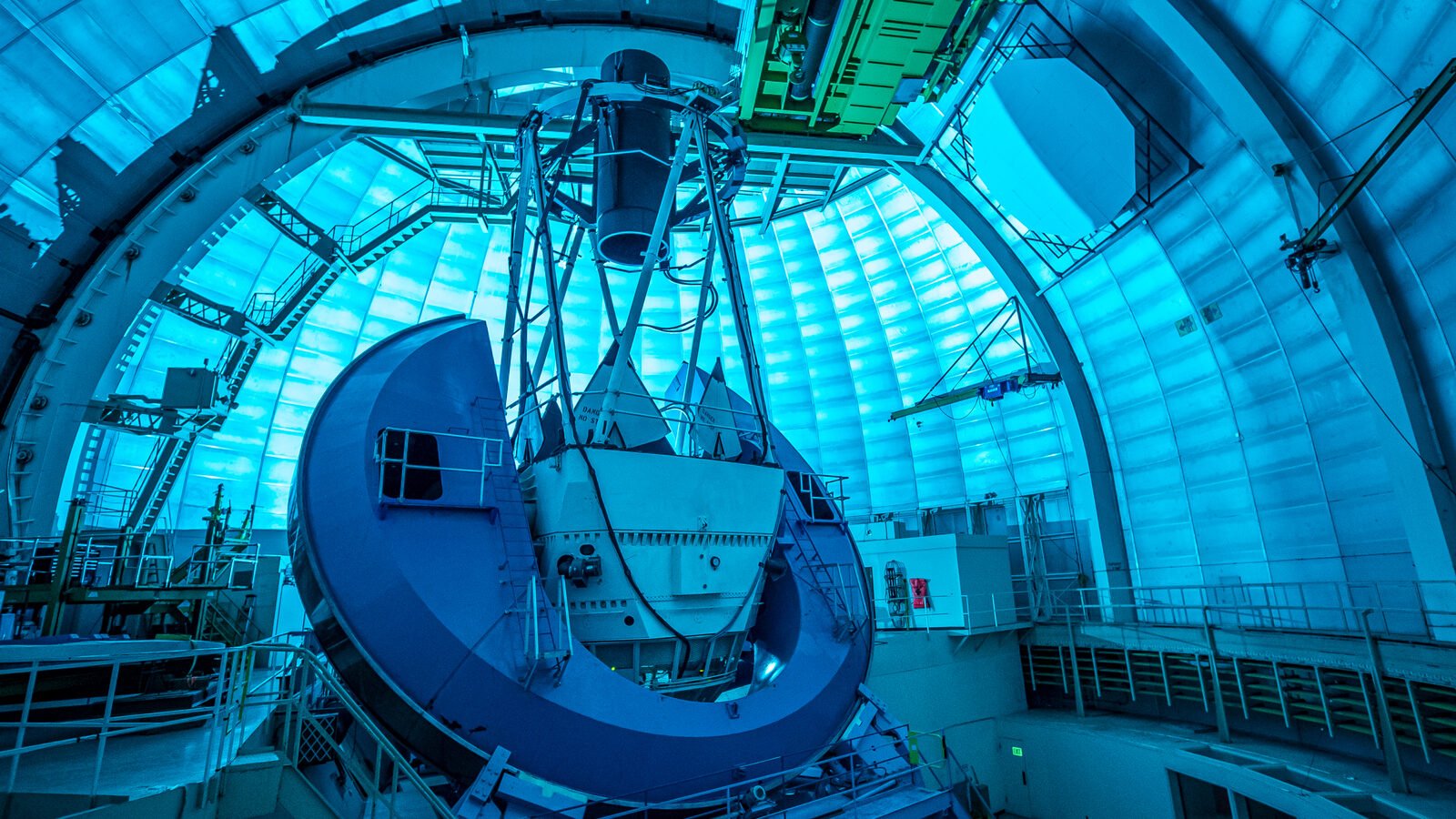 DESI’s instrument, a long black cylinder supported by the metal telescope framework, points at the domed ceiling. The dome interior is bathed in blue light.