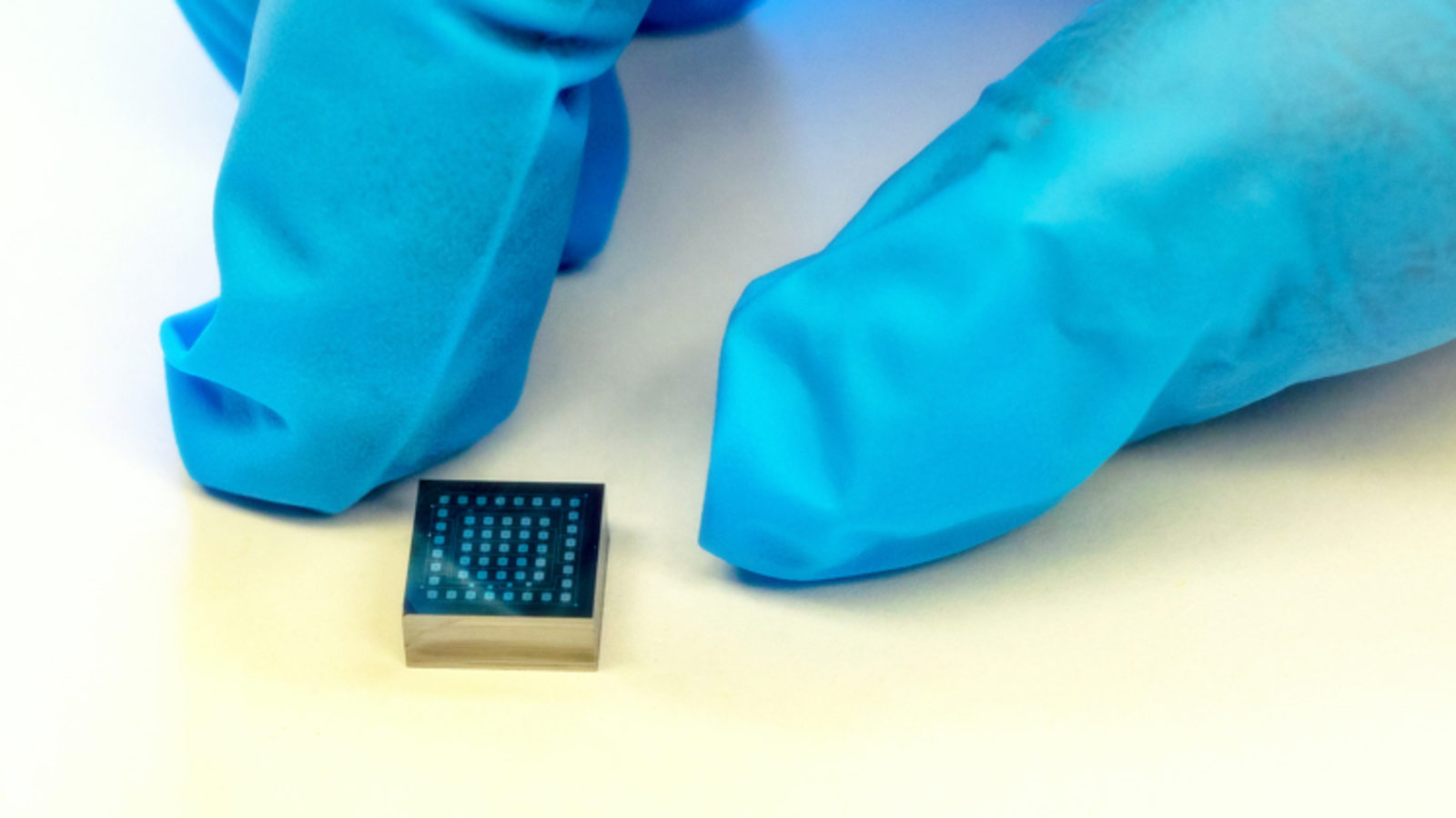 Photo of the index finger and thumb of someone wearing blue gloves next to a cube small enough to fit on a fingertip