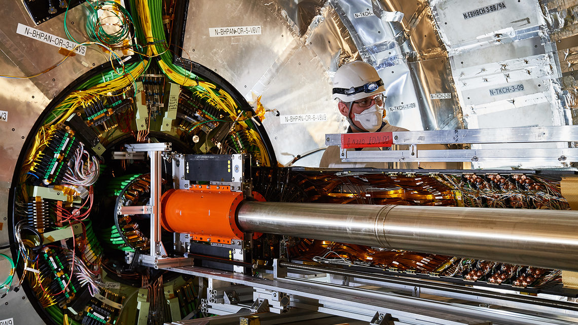 A scientist in cleanroom gear and a hard hat stands by the beam pipe.