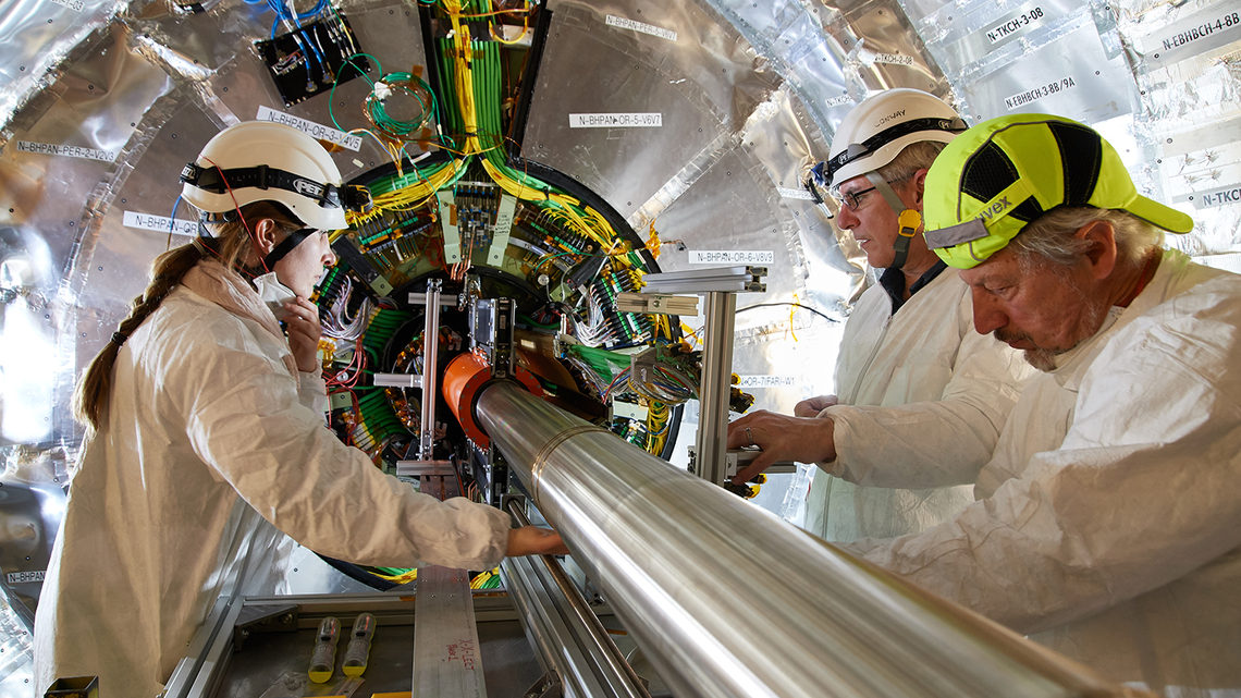 Three scientists in protective helmets talk with their cleanroom masks pulled down or off.