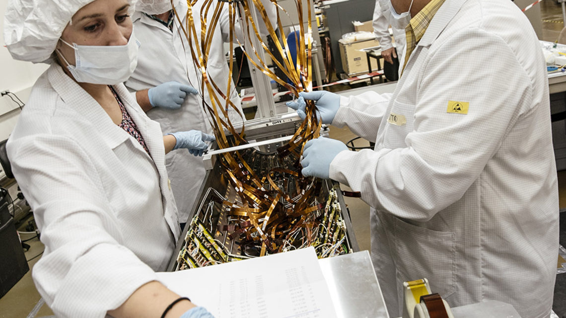 Three scientists in cleanroom garb consult a cabling map.