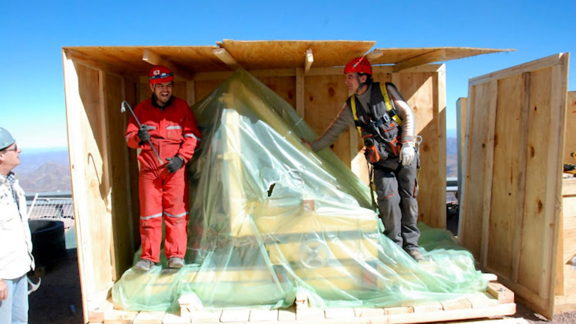 Photo of a support structure for the Dark Energy Camera sits shrouded in plastic while workers disassemble its crate