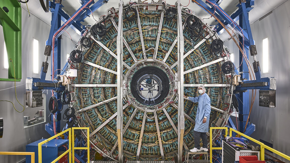 Image of a person in clean-room gear standing in front of a detector component that is twice as tall