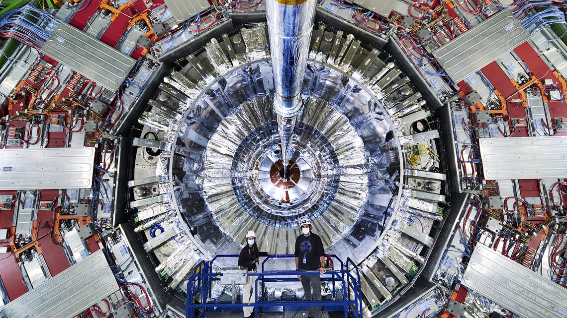 Photo of two people in masks and hard hats standing on a lift next to the CMS detector