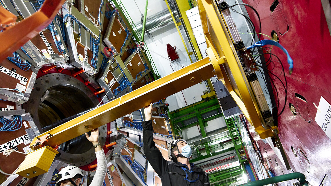 Photo of two people in masks and hard hats on a lift next to the CMS detector