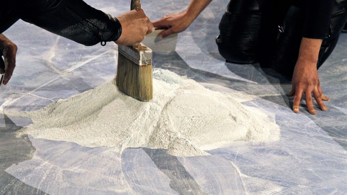 Side view photograph of artists kneeling next to the pile of sand, holding a brush