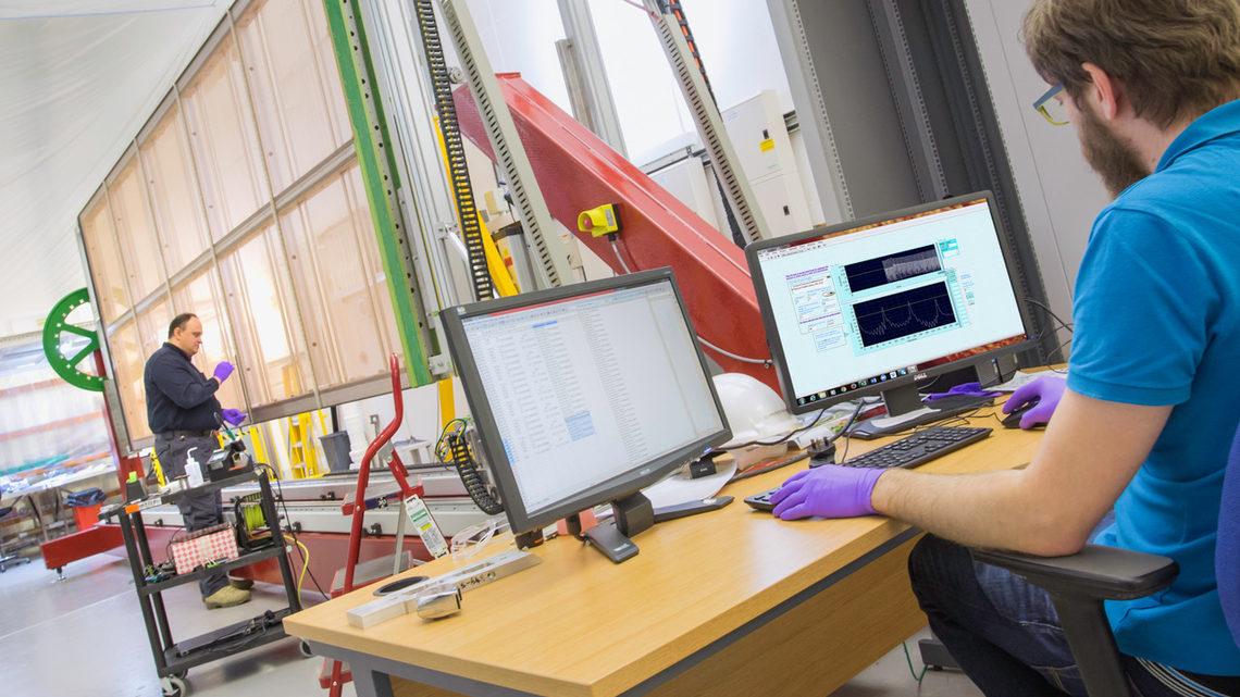 Photo of an individual in purple gloves examining the wires of the APA. A colleague watches a computer monitor