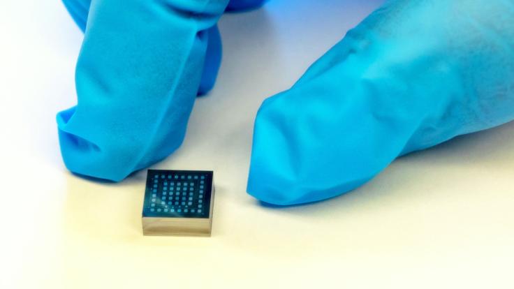 Photo of the index finger and thumb of someone wearing blue gloves next to a cube small enough to fit on a fingertip