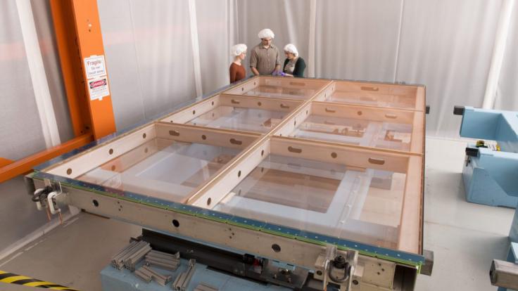 Three physicists with their hair covered stand at the corner of a 4-by-4-meter frame laid flat on a surface