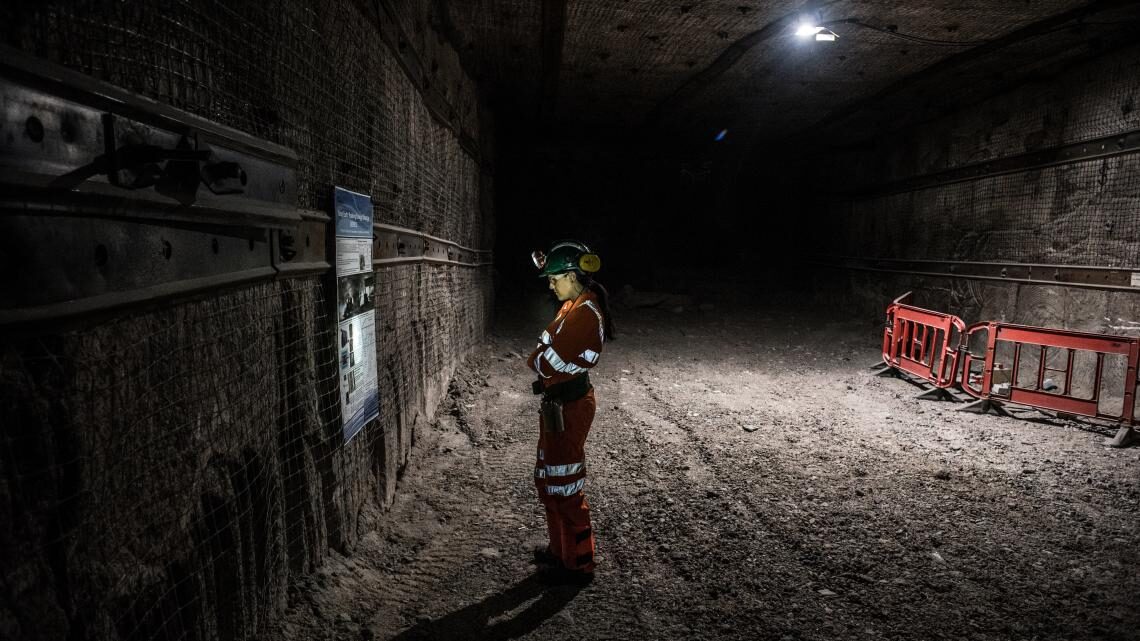 Scientist standing in a mine tunnel, reading a sign, with her face lit by a headlamp on her helmet